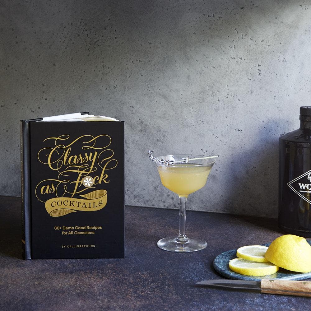 Classy as Fuck Cocktails book with lemon, lavender cocktail and liquor bottle