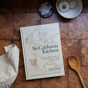 Six California Kitchens with wooden spoon and printed dish towel on a brown tile countertop